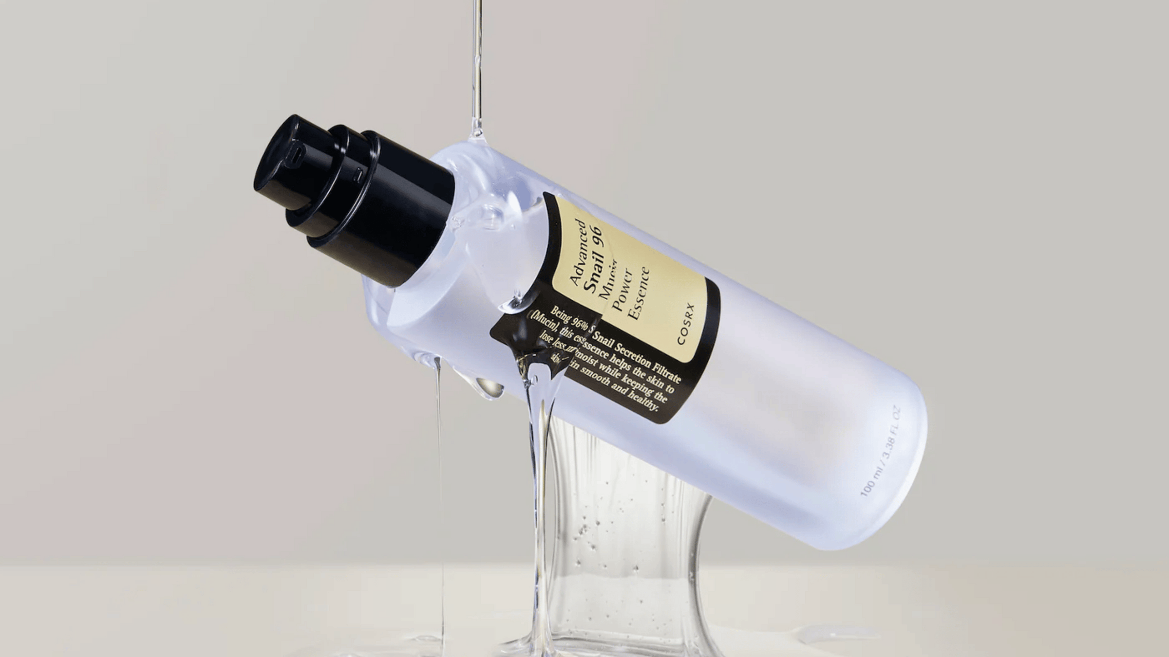 En genomskinlig flaska med COSRX Advanced Snail 96 Mucin Power Essence vält, med essence droppande ner i en glaskärl på en neutral bakgrund.