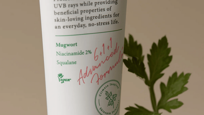 En närbild av en vegansk hudvårdsprodukt med text som lyfter fram ingredienser som mugwort, niacinamid 2 % och squalane. En grön växt visas i bakgrunden, vilket förstärker produktens naturliga och miljövänliga profil.