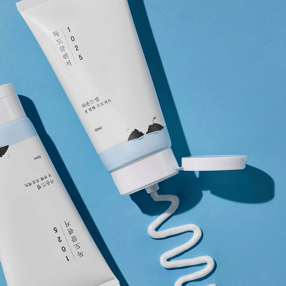 Zwei Tuben von 1025 Dokdo Cleanser auf blauem Hintergrund, eine mit offenem Deckel und einem gewellten Reinigungsschwung herausgedrückt.