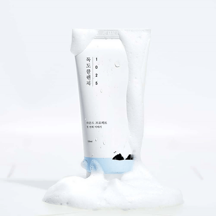 1025 Dokdo Cleanser i vit tub täckt av lödder, med vattenpärlor och skum runt botten, frilagd mot ljus bakgrund.