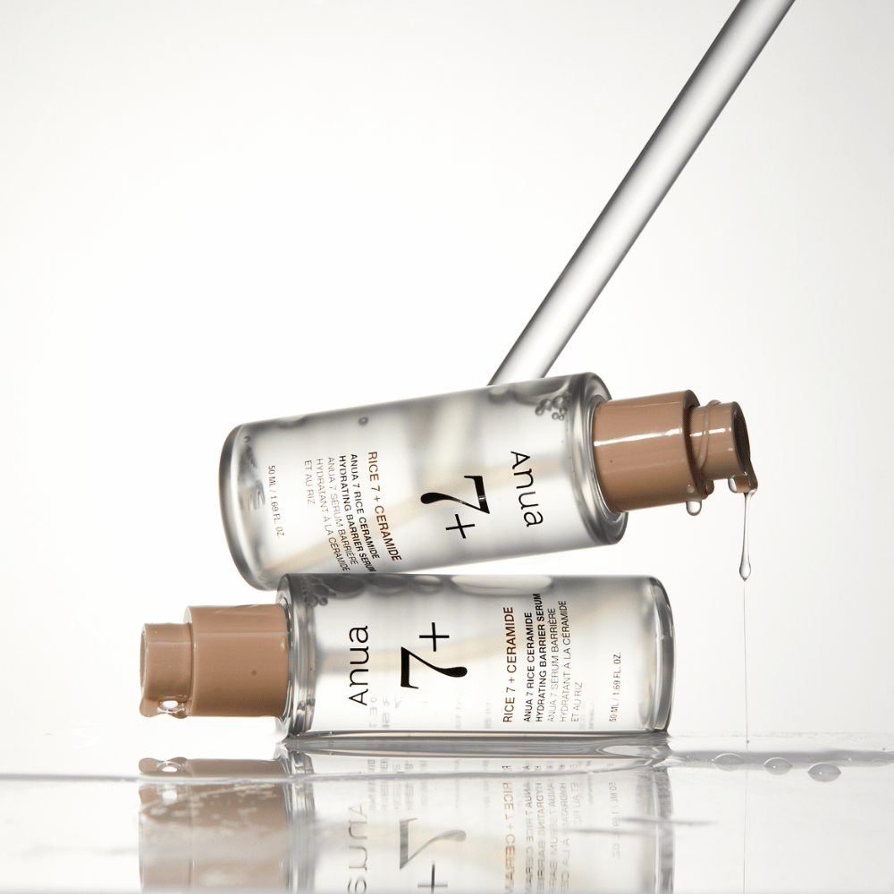 Das Bild zeigt zwei durchsichtige Flaschen des "Anua 77 Rice + Ceramide Hydrating Barrier Serum" mit Tropfen, die von einer Pipette fließen. Das Serum, das mit Reis und Ceramiden angereichert ist, soll die Hautbarriere befeuchten und stärken. Die Flaschen sind elegant gestaltet, was das Premiumgefühl des Produkts verstärkt.
