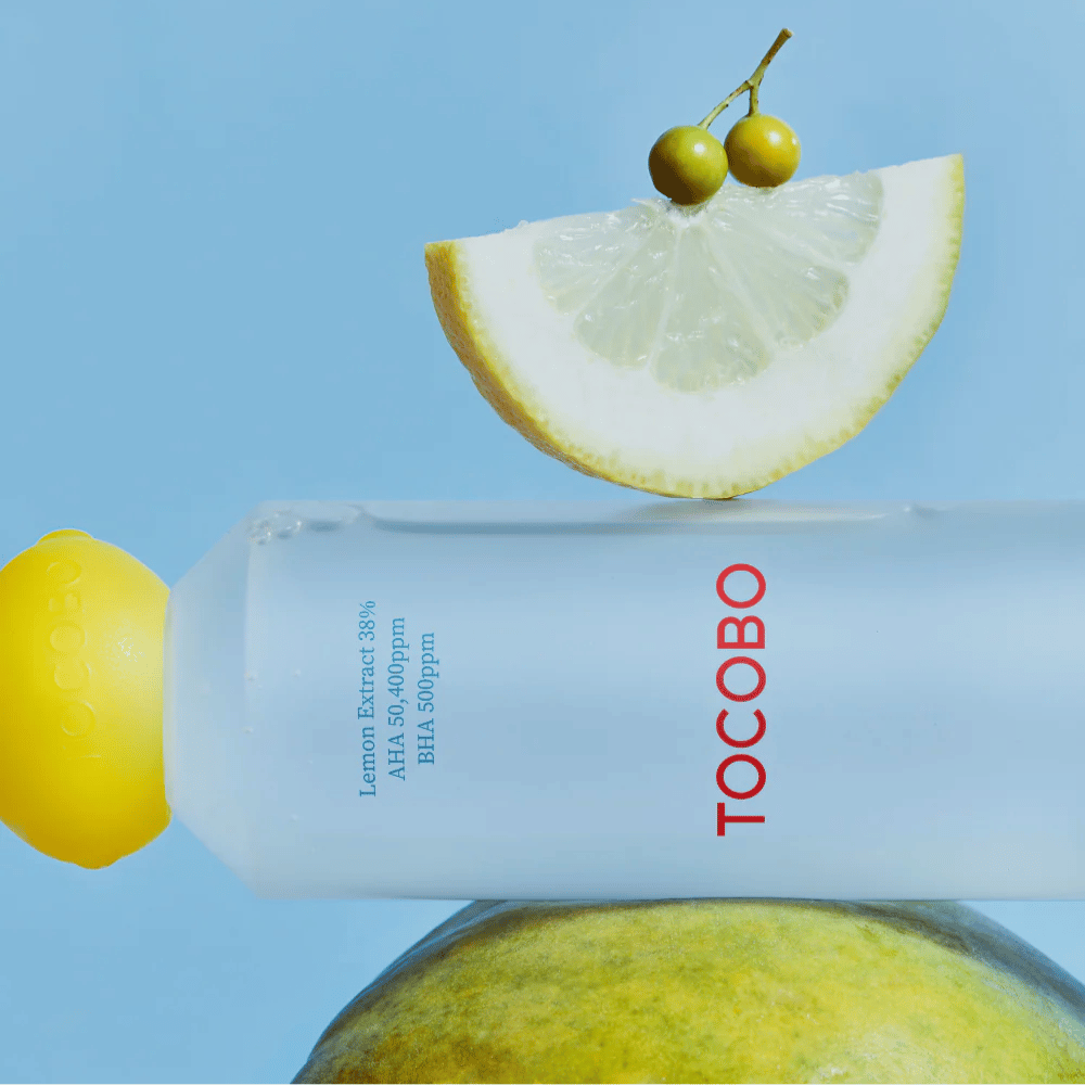 Flasche mit TOCOBO AHA BHA Lemon Toner, verziert mit einem Deckel in Form einer Zitrone. Eine Zitronenscheibe und kleine grüne Beeren balancieren auf der Flasche vor einem blauen Hintergrund.