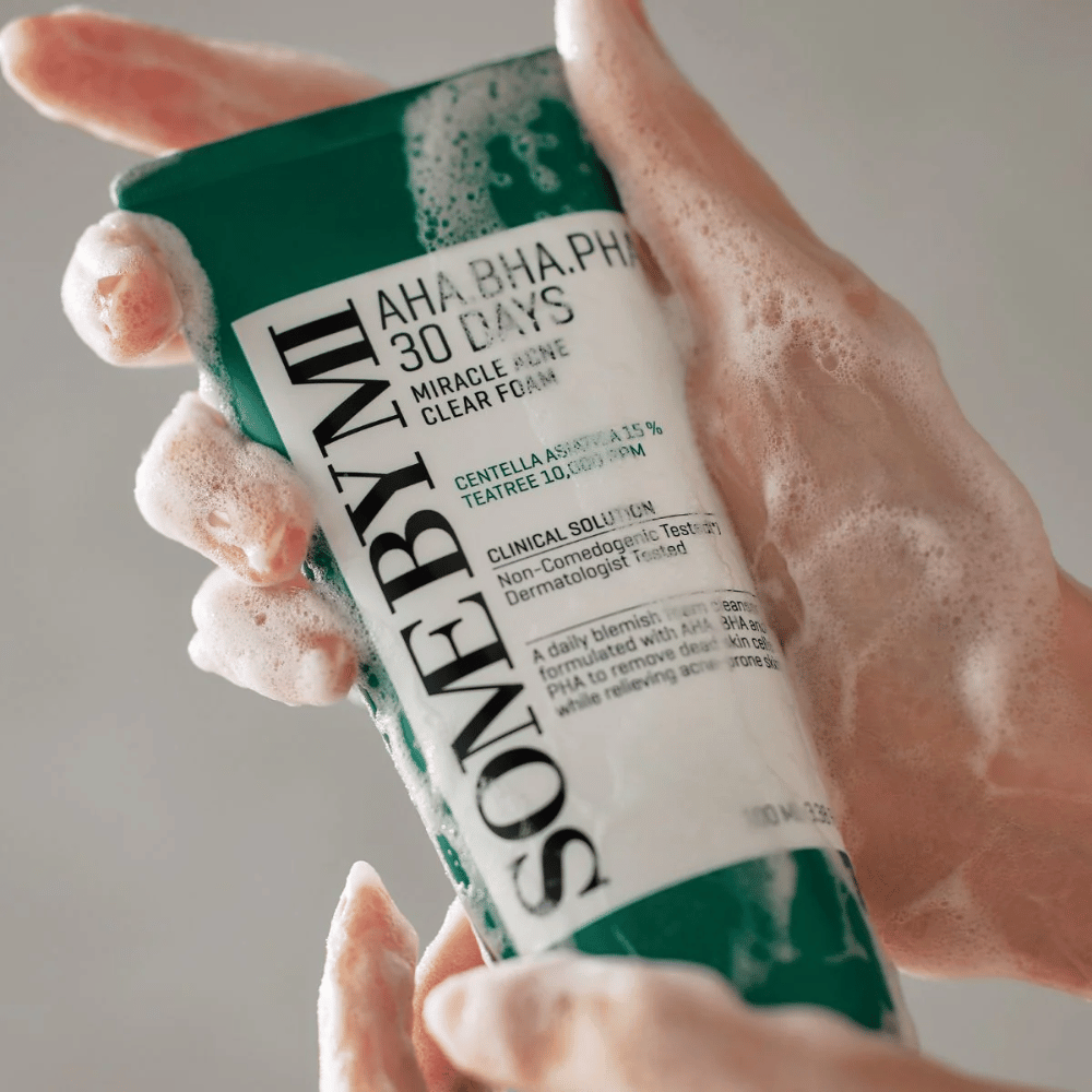 Eine Person hält eine Tube von "SOME BY MI AHA, BHA, PHA 30 Days Miracle Acne Clear Foam" mit schäumendem Inhalt über den Händen. Das Produkt ist für die tägliche Anwendung gedacht, um Akne zu bekämpfen und die Poren mit seinen aktiven Inhaltsstoffen wie Centella, tea tree Öl und Säuren wie AHA, BHA und PHA zu reinigen.