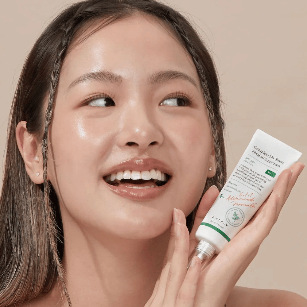 Das Bild zeigt eine junge Frau, die fröhlich lächelt und eine Tube mit AXIS-Y Complete No-Stress Physical Sunscreen hält. Sie hat ein strahlendes und natürliches Aussehen mit minimalem Make-up, was das Produkt, das sie präsentiert, hervorhebt.