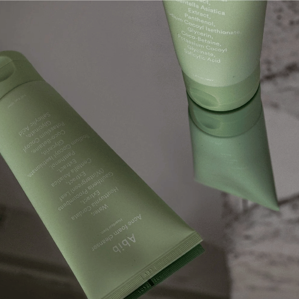 Grüne Tube von Abib Acne Foam Cleanser Heartleaf liegt auf einem Spiegelbord mit der Vorderseite des Produkts und gespiegeltem Text. Zutaten wie Salicylsäure, Centella und Herzblattextrakt sind deutlich auf der Verpackung zu sehen.