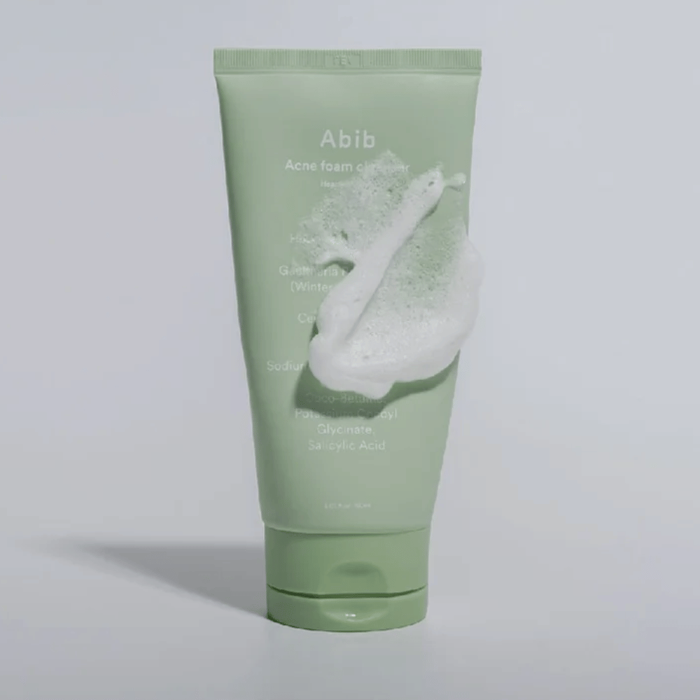 Grüne Tube mit Abib Acne Foam Cleanser Heartleaf, bedeckt von einem weißen Schaum, der den schäumenden Reinigungseffekt illustriert. Das Produkt enthält Herzblattextrakt und Salicylsäure für zu Akne neigende Haut.