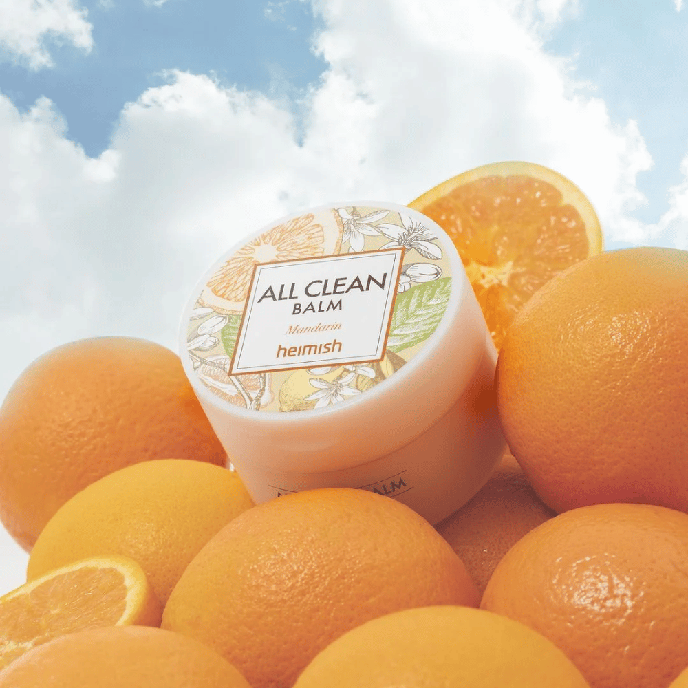 Glas von Heimish All Clean Balm Mandarin auf einem Haufen Orangen platziert, mit einem blauen Himmel im Hintergrund.