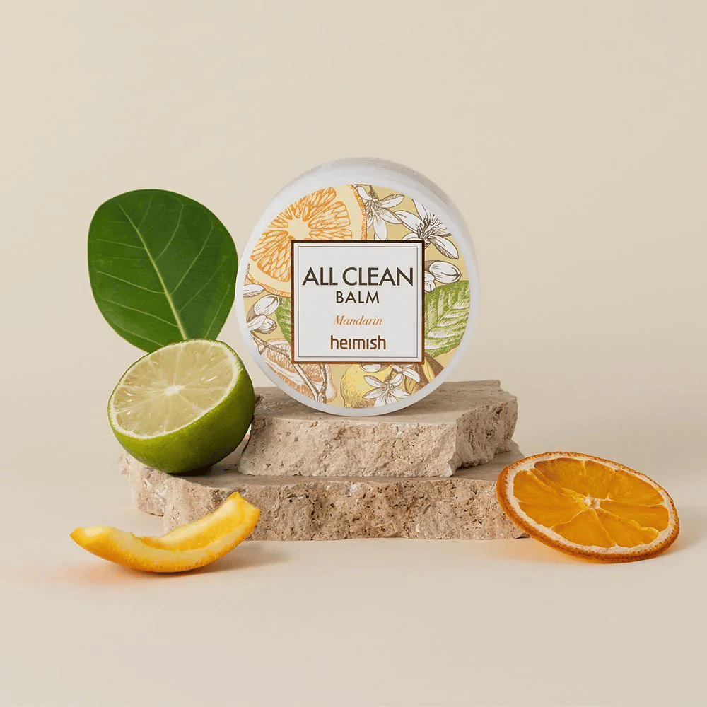 Glas von Heimish All Clean Balm Mandarin auf Steinen platziert, umgeben von Mandarinenschalen und Zitrusblättern, vor einem hellen Hintergrund.