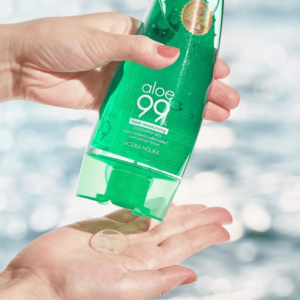 Eine Hand hält eine grüne Tube mit Holika Holika Aloe 99% Soothing Gel, während eine andere Hand einen Klecks Gel auffängt, der ausgegeben wird. Der Hintergrund ist unscharf und zeigt ein wässriges, glitzerndes Muster.