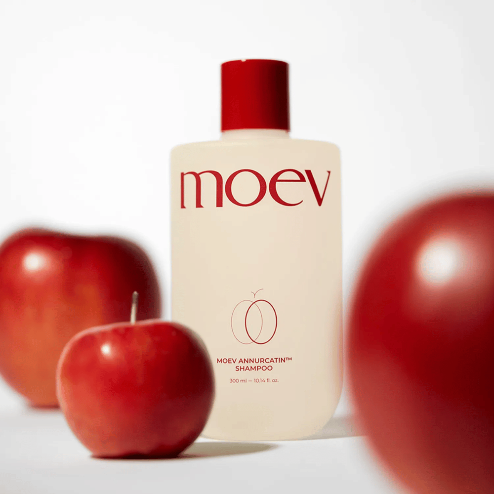 MOEV Annurcatin Shampoo platziert zwischen roten Äpfeln – inspiriert von den Antioxidantien des Annurca-Apfels, die stärkeres, gesünderes und glänzenderes Haar verleihen.