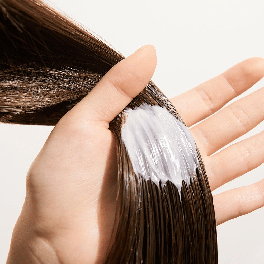 Hand trägt Nature Republic Argan Essential Moisture Repairing Hair Mask auf nasses Haar auf – feuchtigkeitsspendende Maske, die das Haar stärkt und weich macht.