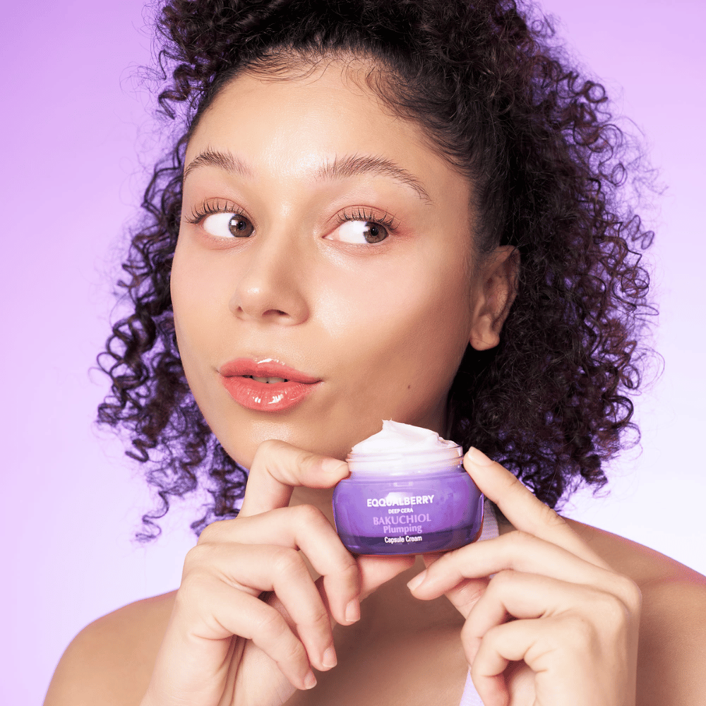 Frau hält EQQUALBERRY Bakuchiol Plumping Capsule Cream in der Hand und zeigt ihre befeuchtete, strahlende Haut vor einem lila Hintergrund.