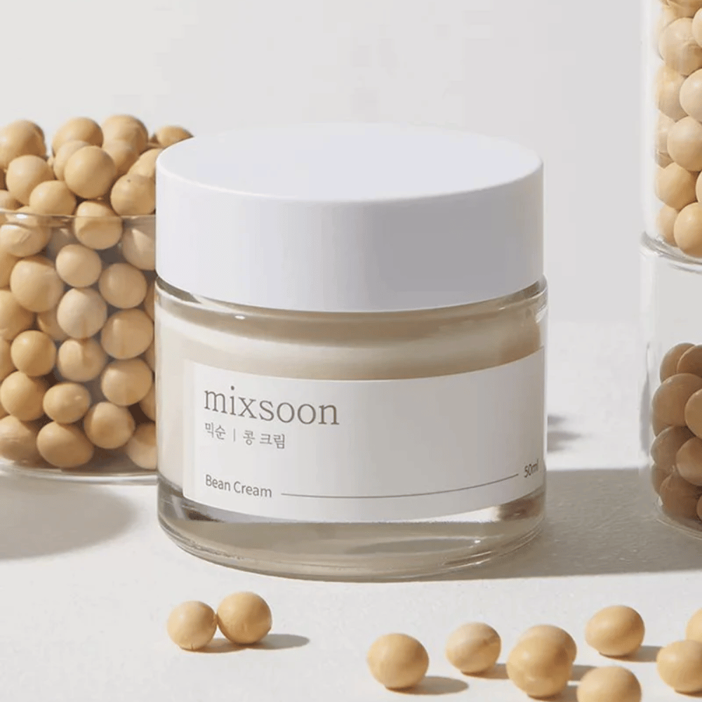 Ein transparenter Behälter mit weißem Etikett und Deckel, etikettiert mit "mixsoon 팥 크림" und "Bean Cream" mit einem angegebenen Volumen von "50ml". Der Behälter enthält eine hellgelbe Creme und steht auf einer hellen Oberfläche. Neben dem Behälter befindet sich ein Glasbehälter voller hellbrauner Bohnen, die auf die Oberfläche verschüttet wurden, was darauf hindeutet, dass die Creme Bohnenextrakt enthält. Der Hintergrund und die Beleuchtung schaffen eine warme und natürliche Atmosphäre.