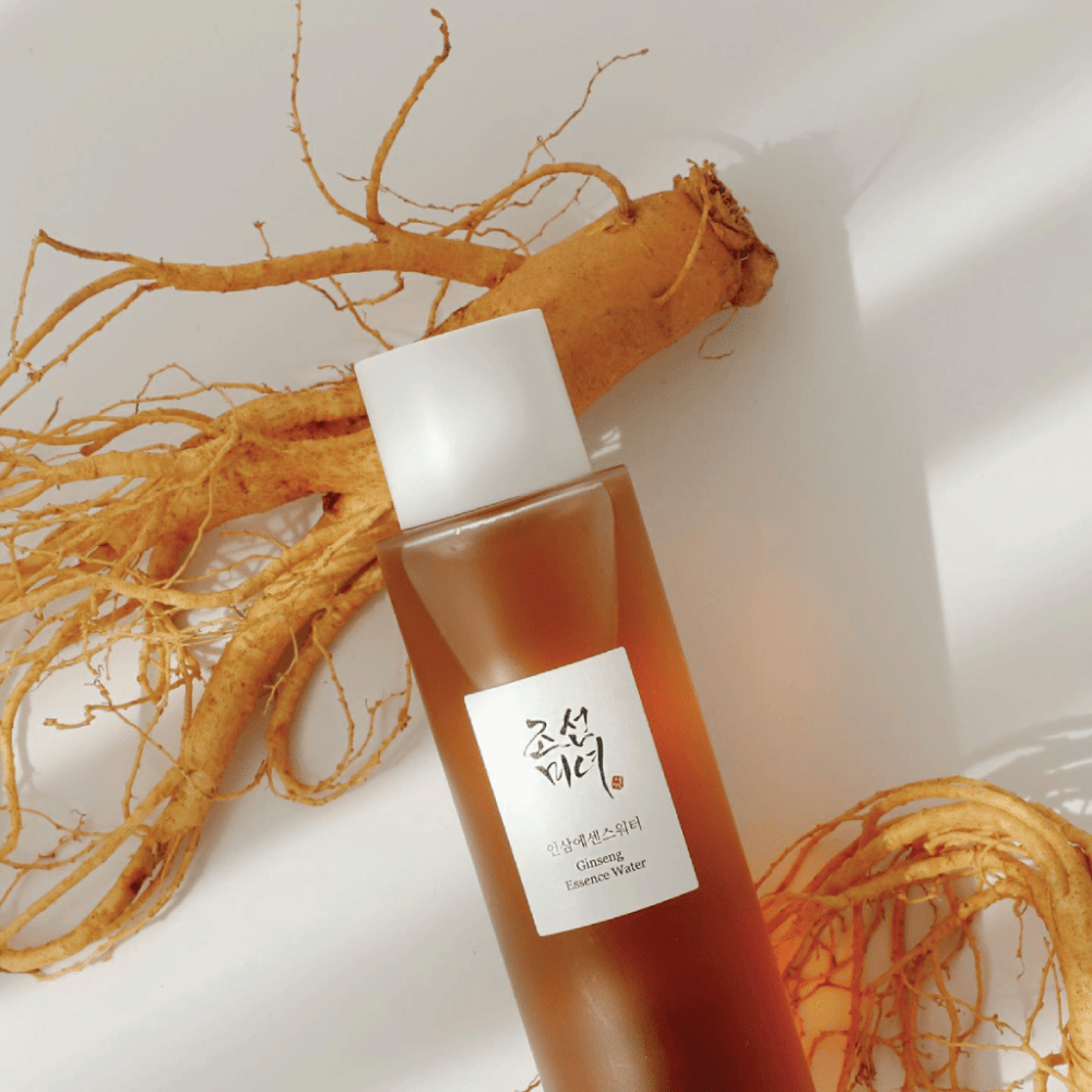 Eine braune Flasche Beauty of Joseon Ginseng Essence Water mit weißem Verschluss liegt neben einer frischen Ginsengwurzel auf weißem Hintergrund.