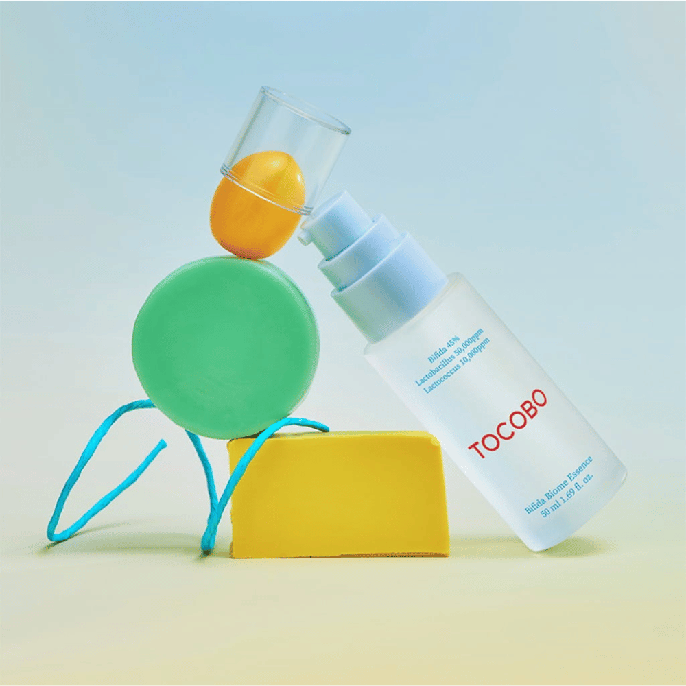 TOCOBO Bifida Biome Essence in mattierter Pumpflasche mit rotem Logo, platziert vor einem farbenfrohen Hintergrund mit grünen, gelben und orangefarbenen geometrischen Formen. Der Text auf der Flasche gibt Bifida 5 %, Lactobacillus 3 000 ppm und Lactococcus 1 000 ppm an.