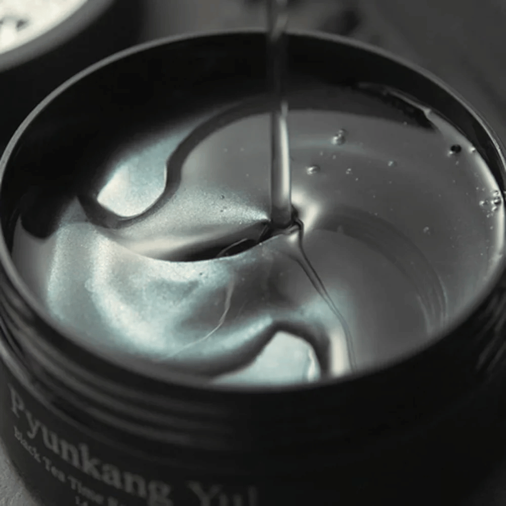 Nahaufnahme von schwarzen Hydrogel-Augenpads in einem geöffneten Tiegel von Pyunkang Yul. Die glänzenden Pads liegen in Serum und unterstreichen das feuchtigkeitsspendende und exklusive Gefühl des Produkts.