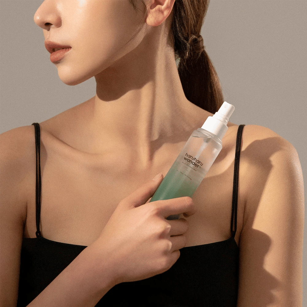 Bild von HaruHaru Wonder Black Bamboo Mist gehalten von einer Frau. Das Produkt befindet sich in einer transparenten Sprühflasche mit einem grünen Ton und einer weißen Pumpe. Die Frau hat einen entspannten Look, trägt ein schwarzes Oberteil und konzentriert sich darauf, das Spray zu verwenden, um der Haut Feuchtigkeit und Glanz zu verleihen. Die Flasche ist Teil einer Hautpflegeroutine.