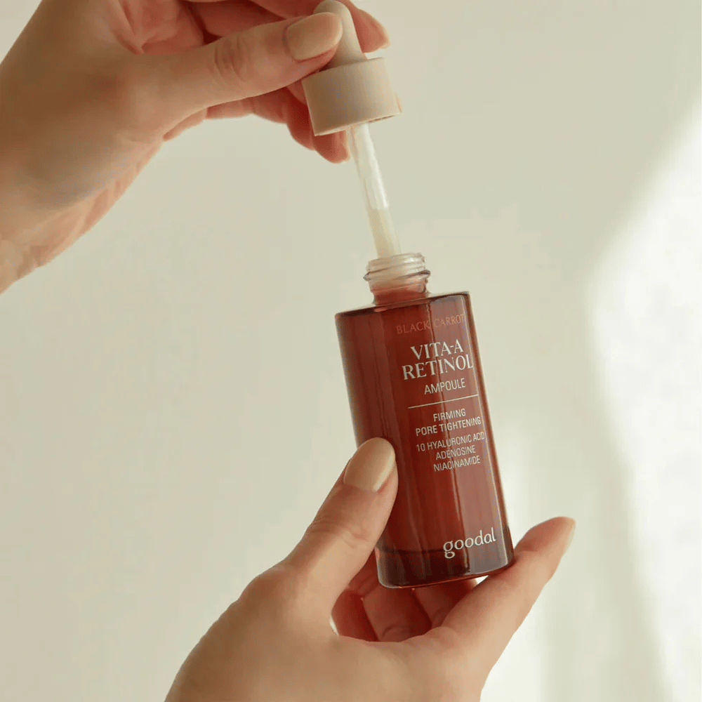 Eine Hand hält eine Flasche mit Goodal Black Carrot Vita-A Retinol Firming Ampoule. Das Flaschenetikett zeigt den Produktnamen und die Eigenschaften, einschließlich "Firming Pore Tightening" und Inhaltsstoffe wie 10% Hyaluronsäure, Adenosin und Niacinamid. Die Pipette ist beige.