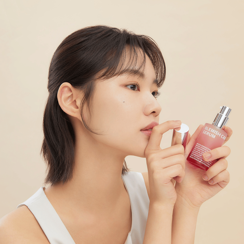 Eine Frau mit dunklem Haar hält eine Flasche Blemish Care Serum in einer Hand und nimmt mit der anderen den Verschluss ab. Sie hat einen neutralen Gesichtsausdruck und der Hintergrund ist hellbeige, was einen weichen und minimalistischen Eindruck vermittelt.