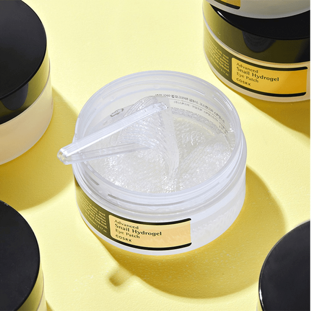 Ein offenes Glas COSRX Advanced Snail Hydrogel Eye Patch mit transparenten Augenpatches und einem kleinen Spatel, umgeben von weiteren geschlossenen Gläsern auf gelbem Hintergrund.