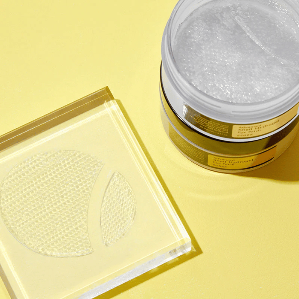 Ein offenes Glas COSRX Advanced Snail Hydrogel Eye Patch steht auf gelbem Hintergrund neben einem transparenten Untersetzer, auf dem zwei Augenpatches liegen.