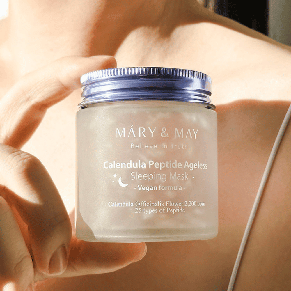 Eine Frau hält ein Glas mit MARY & MAY Calendula Peptide Ageless Sleeping Mask. Das Glas ist transparent mit einem lila Deckel und der Text "Calendula Peptide Ageless Sleeping Mask" ist deutlich sichtbar. Das Produkt ist eine vegane Schlafmaske mit Calendula und Peptiden, die der Haut über Nacht einen jugendlichen Glanz verleihen soll.