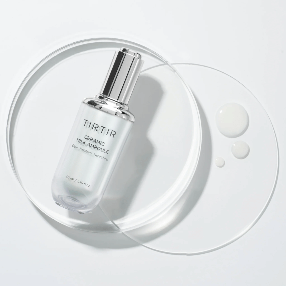 Eine transparente Flasche TIRTIR Ceramic Milk Ampoule auf einer Glasplatte mit Tropfen, vor einem weißen Hintergrund, betont Glanz, Feuchtigkeit und Nährstoffe.