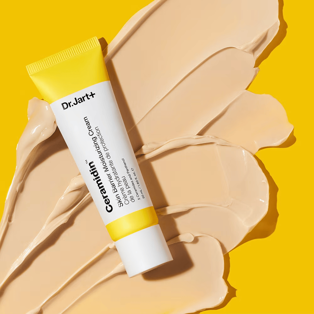 Tube mit Dr. Jart+ Ceramidin Skin Barrier Moisturizing Cream vor einem gelben Hintergrund mit verstreuter Creme.