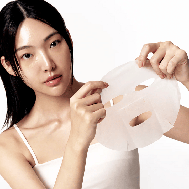 Kvinna med ren och återfuktad hud håller upp en vit sheet mask framför sig, redo att applicera den på ansiktet.