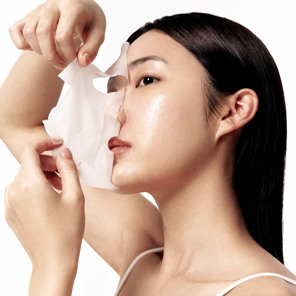 Kvinna med slät, återfuktad hud tar av en vit sheet mask från ansiktet, vilket avslöjar en strålande och jämn hudton.