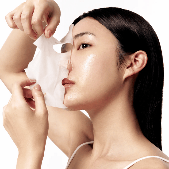 Kvinna med slät, återfuktad hud tar av en vit sheet mask från ansiktet, vilket avslöjar en strålande och jämn hudton.