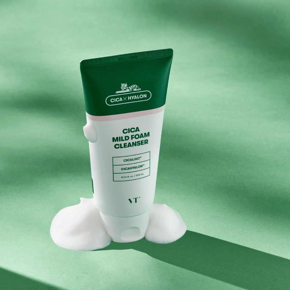Tube mit VT Cica Mild Foam Cleanser stehend auf einem grünen Untergrund mit Schaum um die Basis. Das Produkt hat ein grünes und weißes Design und wird mit CICALIAO™ und CICAHYALON™ vermarktet.