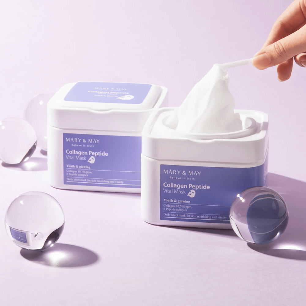 Eine Hand nimmt eine Maske aus einem Behälter von MARY & MAY Collagen Peptide Vital Mask. Die Maske ist für die tägliche Anwendung gedacht, um der Haut Nährstoffe und Glanz zu verleihen, mit Kollagen und Peptiden zur Unterstützung der Jugendlichkeit der Haut.