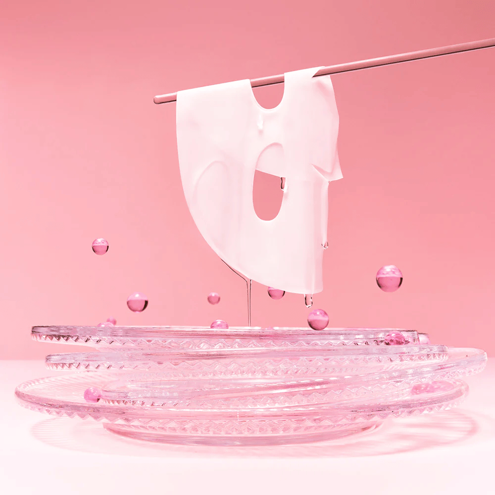 Eine Hydrogel-Maske hängt an einer Stange, tropft mit Serum. Der Hintergrund ist rosa und der Spiegel am Boden reflektiert die rosa Blasen, die die Maske umgeben.