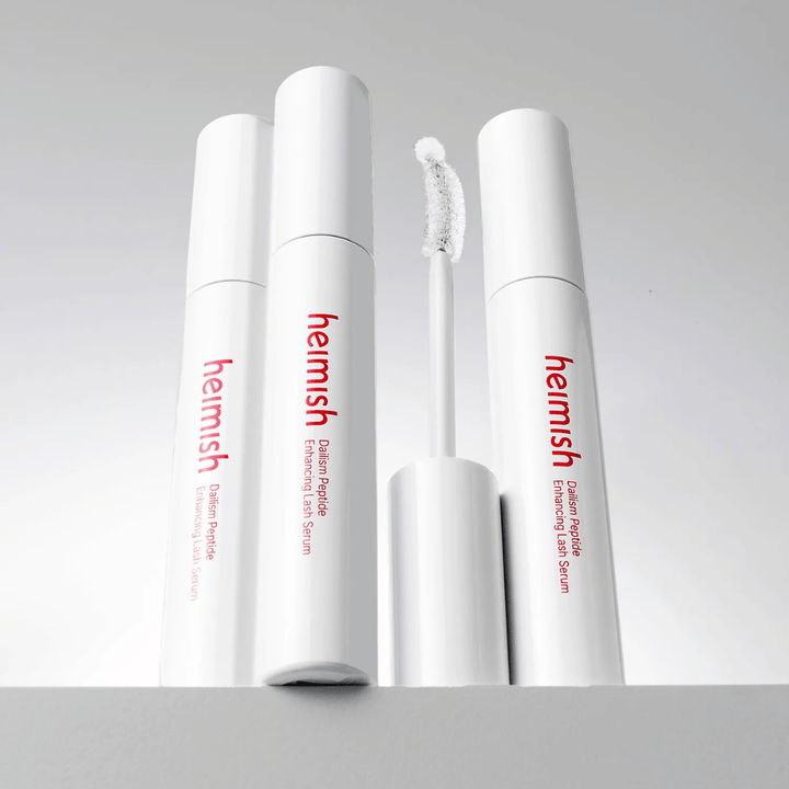 Mehrere Verpackungen von Heimish Dailism Peptide Enhancing Lash Serum werden zusammen mit der gebogenen Applikatorbürste gezeigt. Das Bild hebt das elegante Design des Produkts und die Präzisionsbürste für ein einfaches Auftragen hervor.