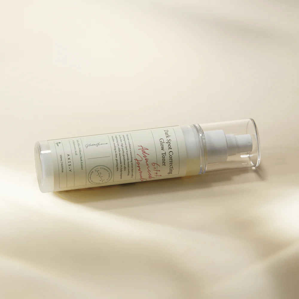 Eine liegende Flasche von "AXIS-Y Dark Spot Correcting Glow Toner" auf einer hellen Oberfläche platziert. Die Flasche ist durchsichtig mit einem weißen Etikett und hat eine weiße Sprühpumpe. Das Etikett zeigt Informationen über die aufhellenden und feuchtigkeitsspendenden Eigenschaften des Produkts und enthält Schlüsselzutaten wie Glutathion und Niacinamid.