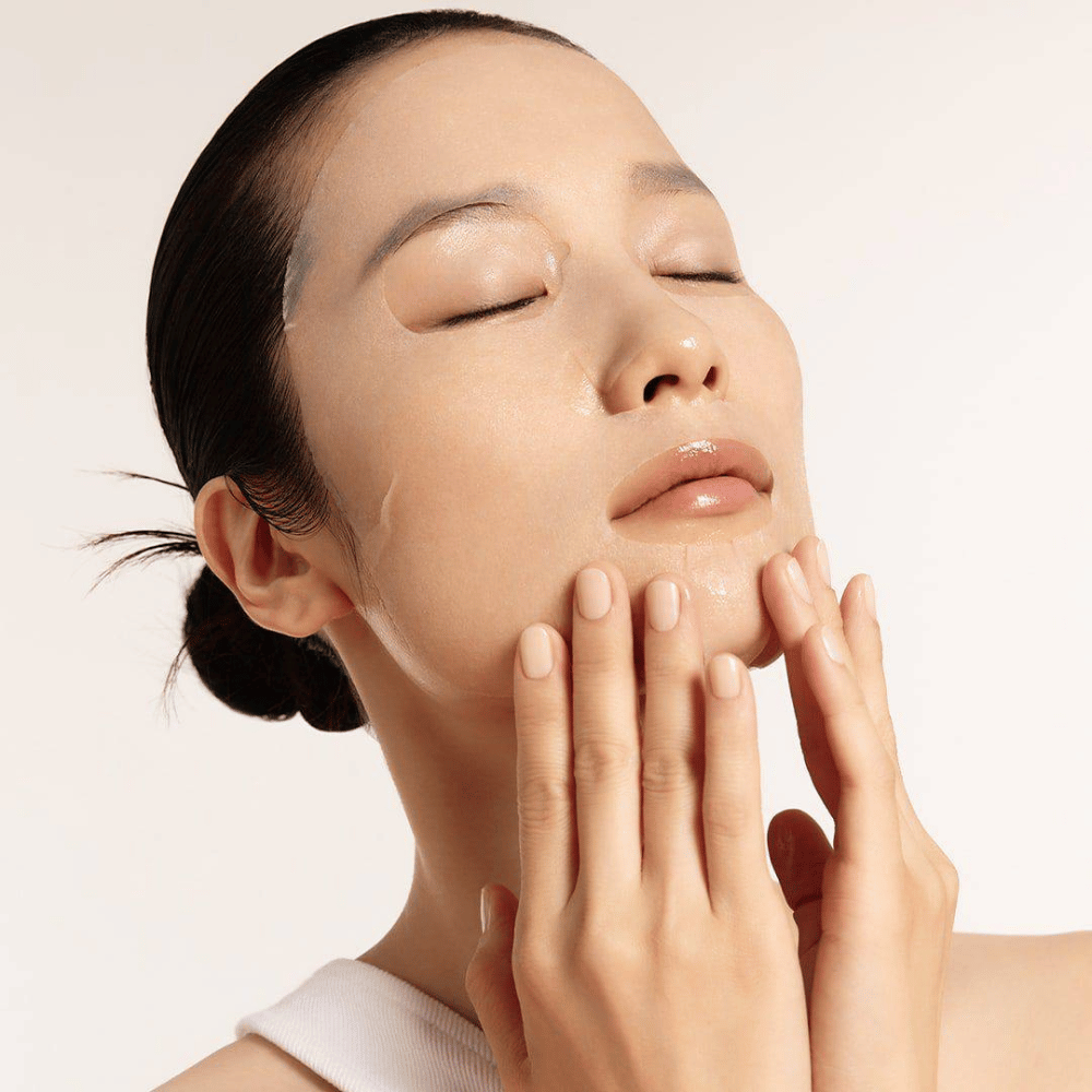 Modell mit hydratisierter und strahlender Haut trägt Sheet mask auf, Fokus auf Strahlkraft, gleichmäßigem Hautton und Glow.