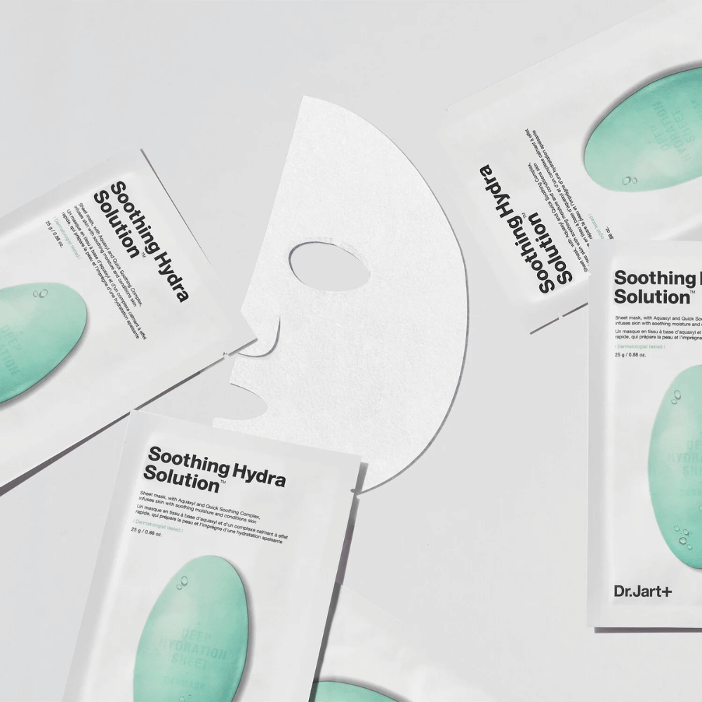 Mehrere Packungen der "Soothing Hydra Solution" Gesichtsmasken von Dr.Jart+ sind auf einer hellen Fläche verstreut. Eine entfaltete Maske ist in der Mitte platziert, welche ihre weiße und perforierte Struktur zeigt.