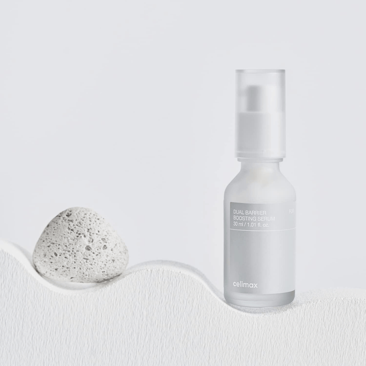 Celimax Dual Barrier Boosting Serum auf Textil mit Stein platziert, ruhiges Produktbild, das Feuchtigkeit und gestärkte Hautbarriere signalisiert.