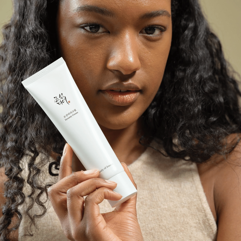 Eine Frau mit lockigem Haar hält eine Tube "Beauty of Joseon Dynasty Cream" nah an ihr Gesicht, um das Produkt hervorzuheben. Die Creme, präsentiert in einer eleganten weißen Tube, ist darauf ausgelegt, die Haut zu nähren und zu verjüngen, was ihre Attraktivität für verschiedene Hauttypen unterstreicht.