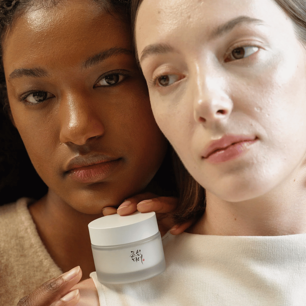 Zwei Frauen, eine mit dunklerem und eine mit hellerer Haut, stehen nah beieinander mit Fokus auf ihren Gesichtern und einer Hautcreme von "Beauty of Joseon". Die Creme ist vor ihnen platziert, was ihre Anwendung für verschiedene Hauttypen und ihre Fähigkeit, Glanz und Nährstoffe zu spenden, betont.
