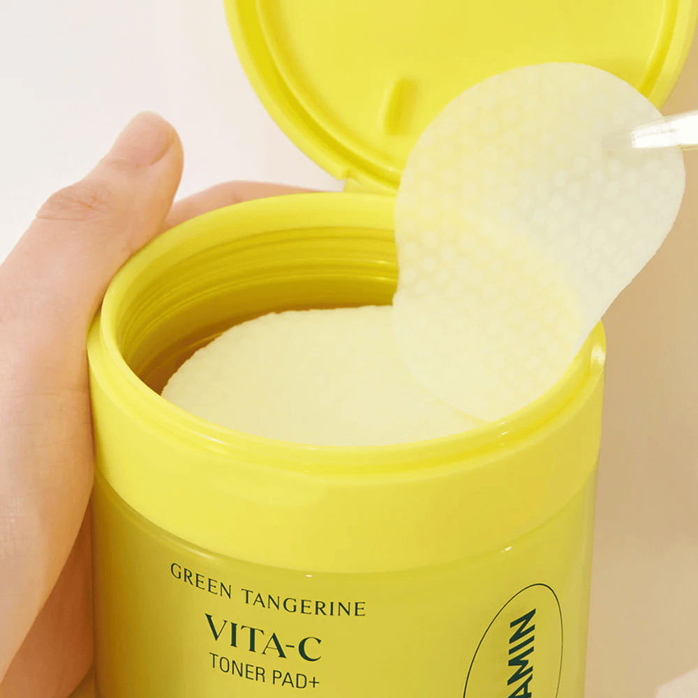 Eine Hand benutzt eine Plastikpinzette, um ein strukturiertes Tonerpad aus einem hellgelben Behälter mit der Aufschrift "GREEN TANGERINE VITA-C TONER PAD+" der Marke "gooda" zu entnehmen.