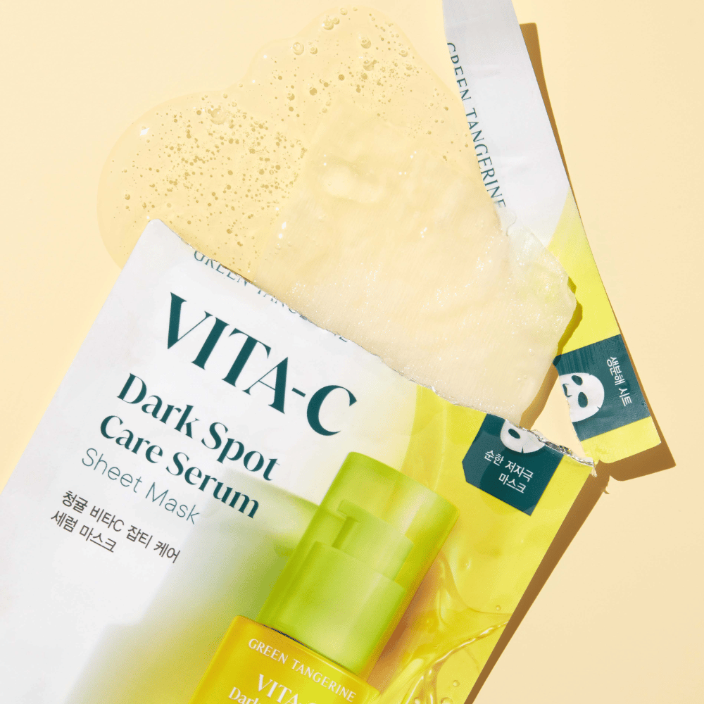 Geöffnete Verpackung des VITA-C Dark Spot Care Serum Sheet mask mit einer Gesichtsmaske darauf und einer grünen Serumflasche, alle vor einem gelben Hintergrund.