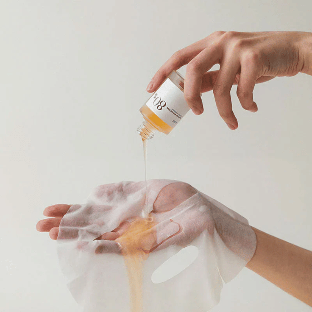 Eine Hand gießt eine goldene, dicke Flüssigkeit aus einer kleinen Flasche, die mit '108' gekennzeichnet ist, auf eine transparente Gesichtsmaske, die in einer anderen Hand gehalten wird. Der Hintergrund ist neutral und einheitlich, um das Produkt hervorzuheben.