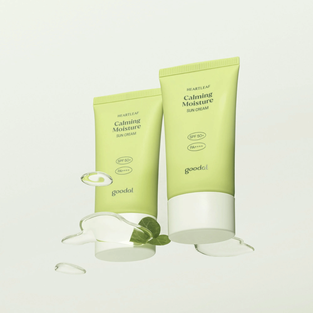 Zwei Tuben von Goodal Heartleaf Calming Moisture Sun Cream SPF50+ PA++++ umgeben von Pflanzenblättern und Serumstropfen vor hellem Hintergrund.