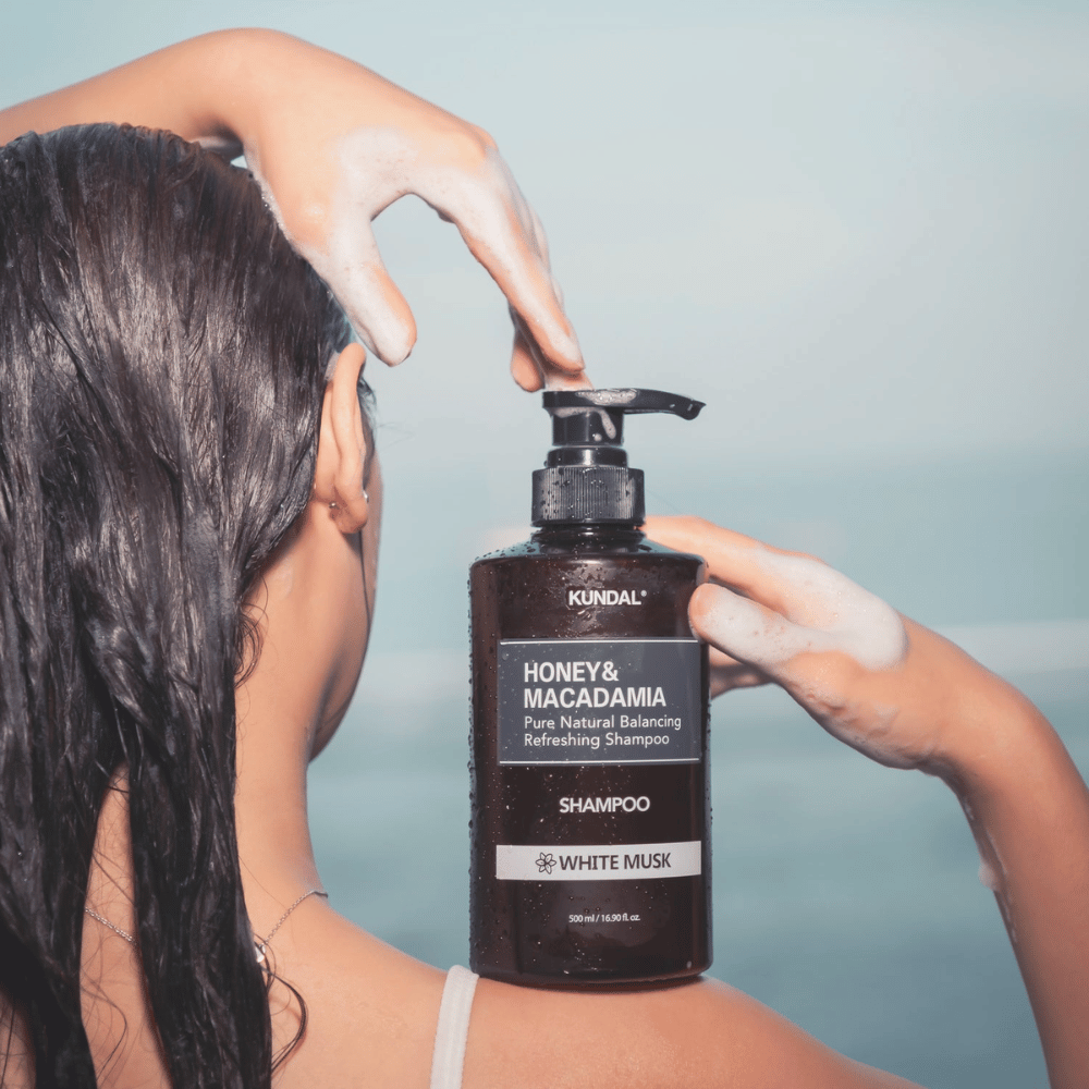 Frau mit nassem Haar trägt Schaum auf und hält KUNDAL Honey & Macadamia Nature Shampoo White Musk vor Meereskulisse.