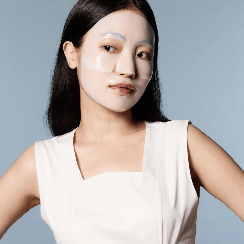 Eine Frau in einem weißen ärmellosen Kleid trägt eine durchsichtige Gesichtsmaske, die fast ihr ganzes Gesicht bedeckt. Die Maske vermittelt den Eindruck fortschrittlicher Hautpflege und gibt das Aussehen einer Schönheitsbehandlung, die darauf ausgelegt ist, die Haut tiefgehend zu befeuchten und zu behandeln.