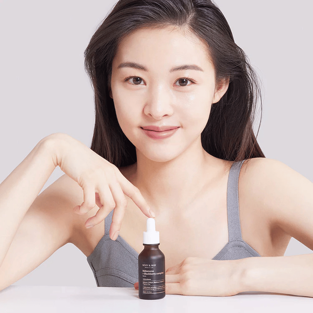 Eine Frau mit langem, dunklem Haar hält MARY & MAY Idebenone + Blackberry Complex Serum, eine dunkle Glasflasche mit Pipette. Sie hat ein fröhliches Lächeln und zeigt mit ihrer anderen Hand auf die Flasche. Die Frau ist in einem grauen, ärmellosen Top gekleidet und scheint sich in einer hellen und einfachen Umgebung zu befinden, was den Fokus des Produkts auf Hautpflege und Schönheit unterstreicht.