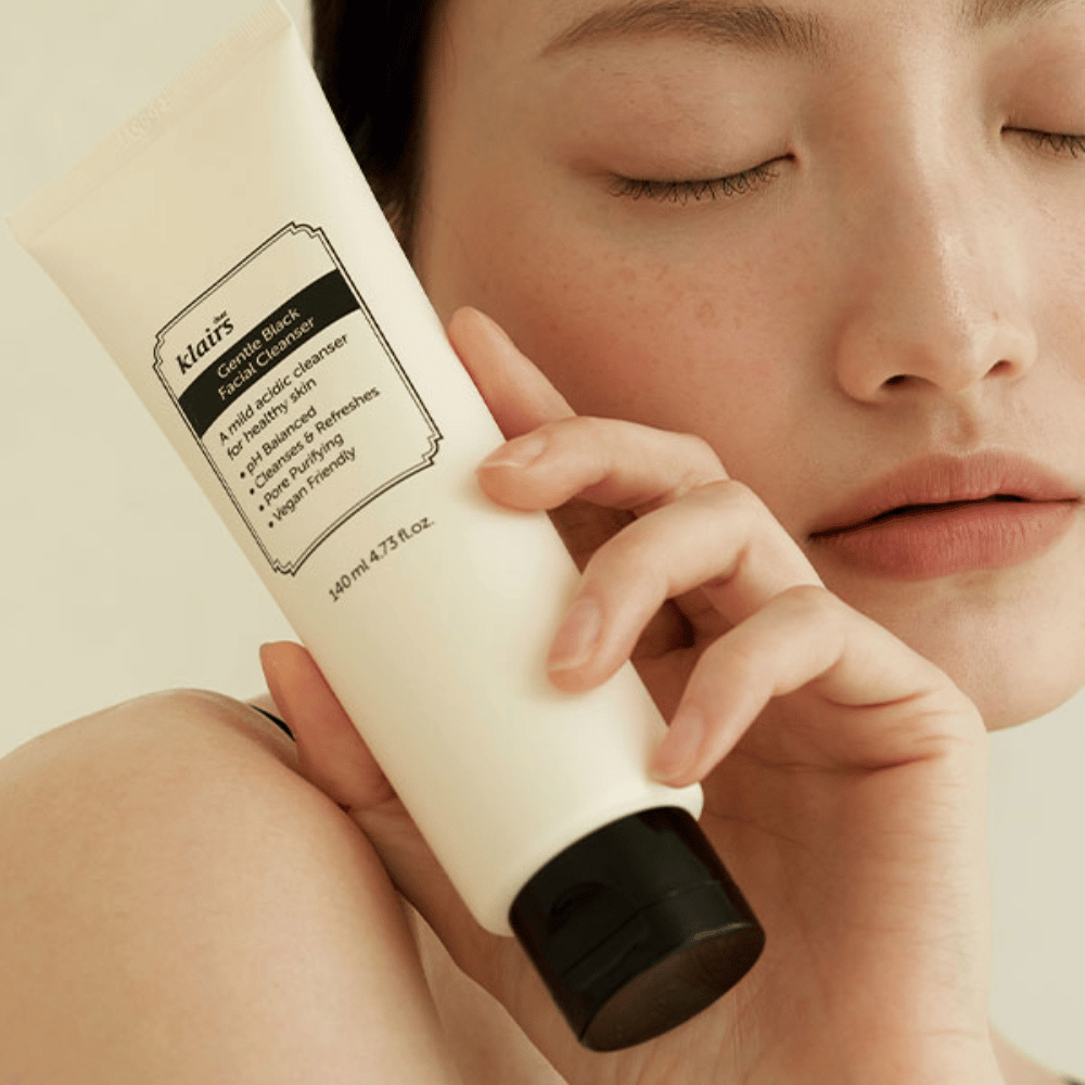 Eine Person hält eine Tube von Klairs Gentle Black Facial Cleanser an ihre Wange, geschlossene Augen und natürlicher Hautton sind im Hintergrund sichtbar.