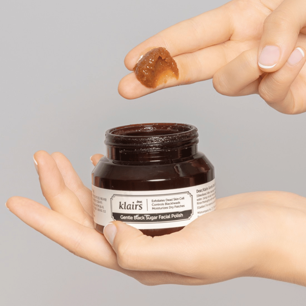 Hände halten eine offene Dose von Klairs Gentle Black Sugar Facial Polish und zeigen das Produkt auf einem Finger, vor einem grauen Hintergrund.